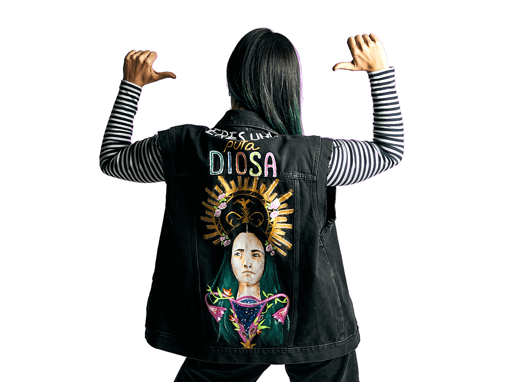 Persona de espaldas, señalando la parte trasera de su chaqueta que tiene bordada la imagen de una mujer con un halo y las palabras "Eres Una Pura Diosa" encima.