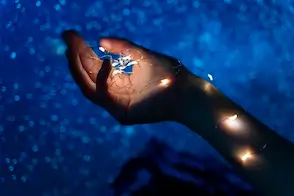 Una mano se extiende sobre un fondo azul estrellado, enérgicamente adornada con pequeñas luces brillantes que la envuelven. La escena evoca una atmósfera mágica y onírica.