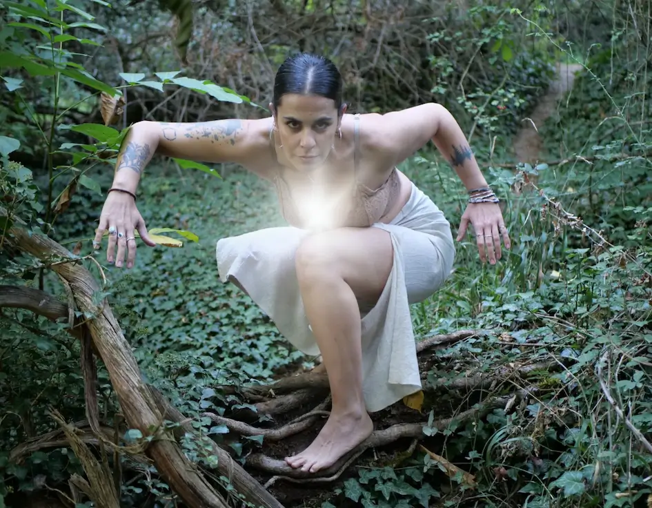 Una persona con tatuajes se agacha descalza sobre ramas retorcidas en un bosque, mirando intensamente a la cámara, con una luz blanca resplandeciente que emana de su pecho.