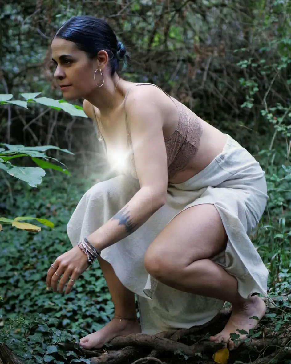 Una mujer con el pelo oscuro recogido en un moño se agacha descalza en un frondoso bosque verde, vestida con una falda beige y un top sin mangas. En su pecho brilla una luz resplandeciente.