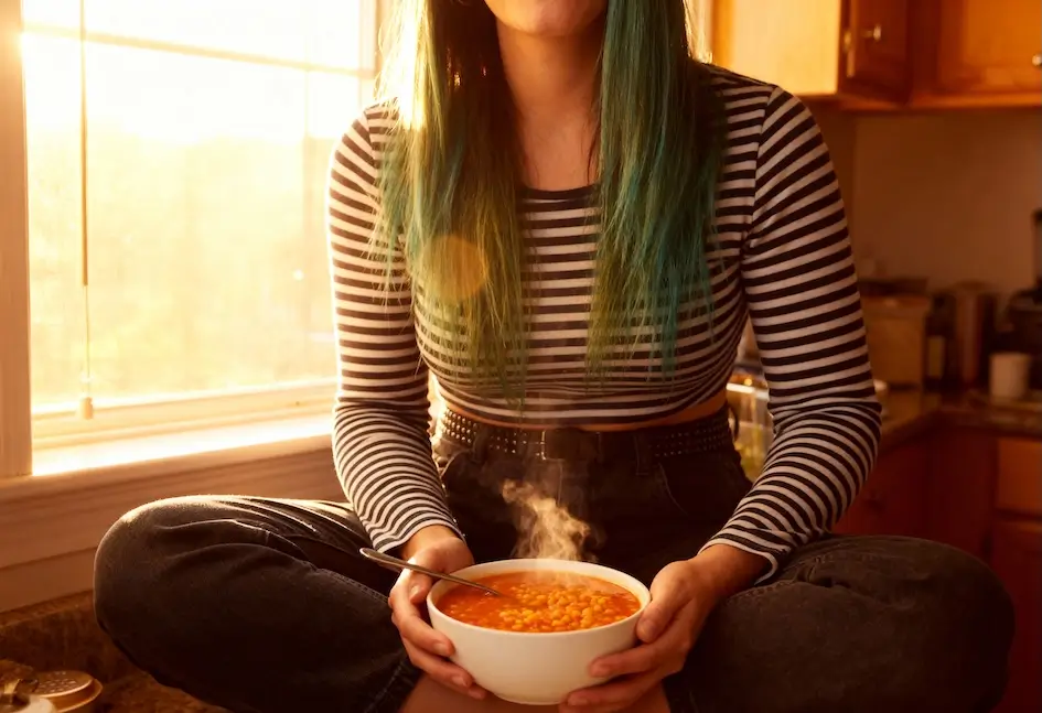 Una persona con el pelo largo y teñido de verde y una camisa de rayas blancas y negras se sienta con las piernas cruzadas en la encimera de la cocina, sosteniendo un humeante plato de sopa, mientras la cálida luz del sol entra por una ventana.
